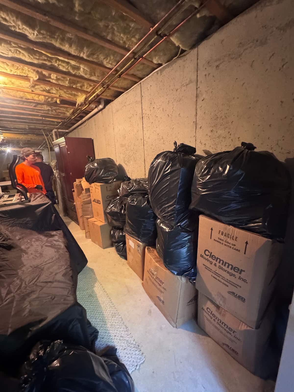 A Middletown basement with full trash bags and moving boxes lined along the wall and two Junk Away crew members working at the far end