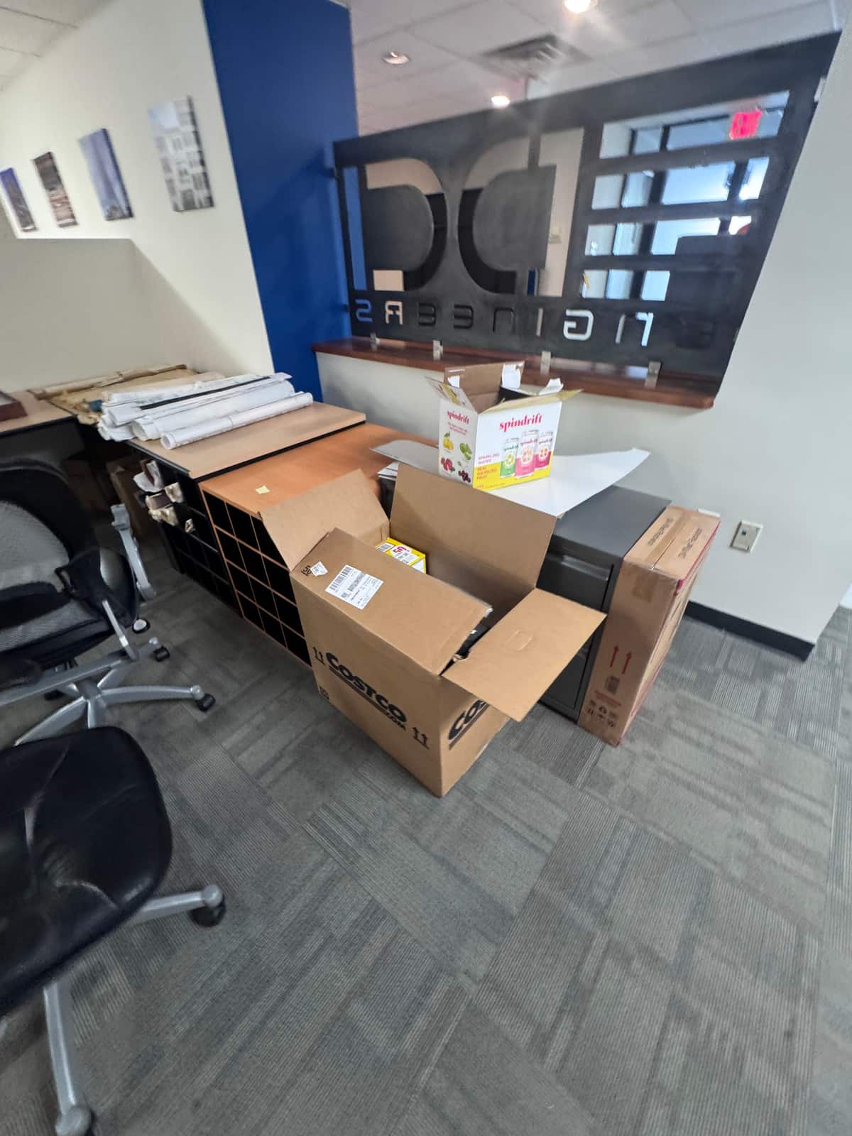 Packed boxes, a cubby shelf unit, a file cabinet and rolled drawings staged in a Wilmington office during a renovation, with desk chairs pulled aside