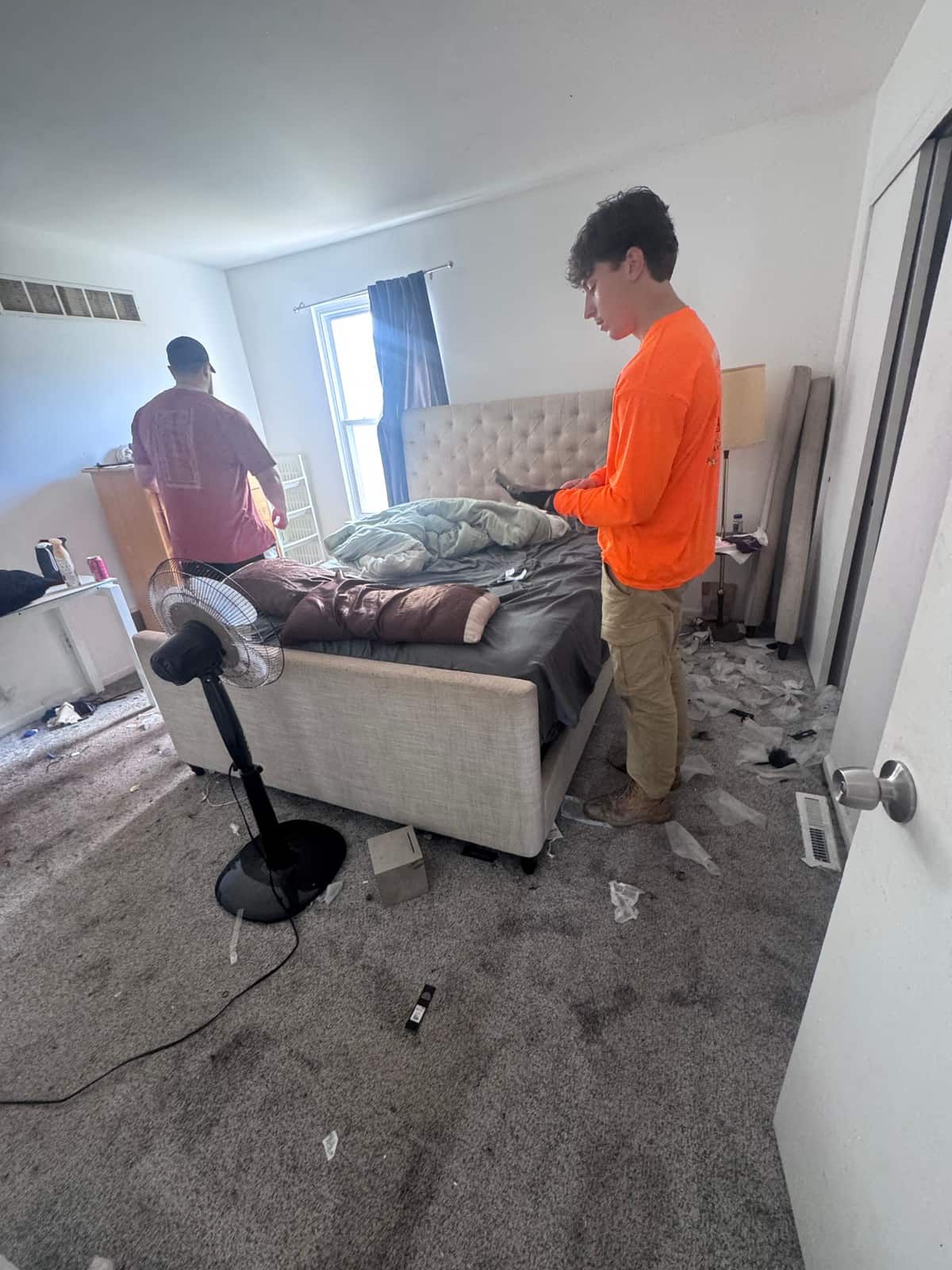 A Newark townhouse bedroom before the cleanout, with a bed, a fan and belongings across the carpet and a Junk Away crew member at work